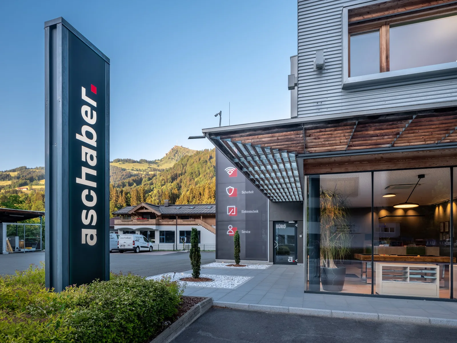 Eingang mit der Aufschrift Büro und der Stele aschaber, im Hintergrund die Berge über Kitzbühel