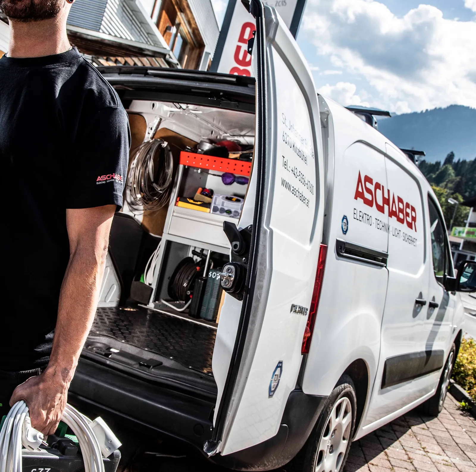 Monteur mit Verlängerungskabel am offenen Firmenbus mit Werkzeugausbau, Beschriftung Aschaber Elektro, Technik, Licht, Sicherheit