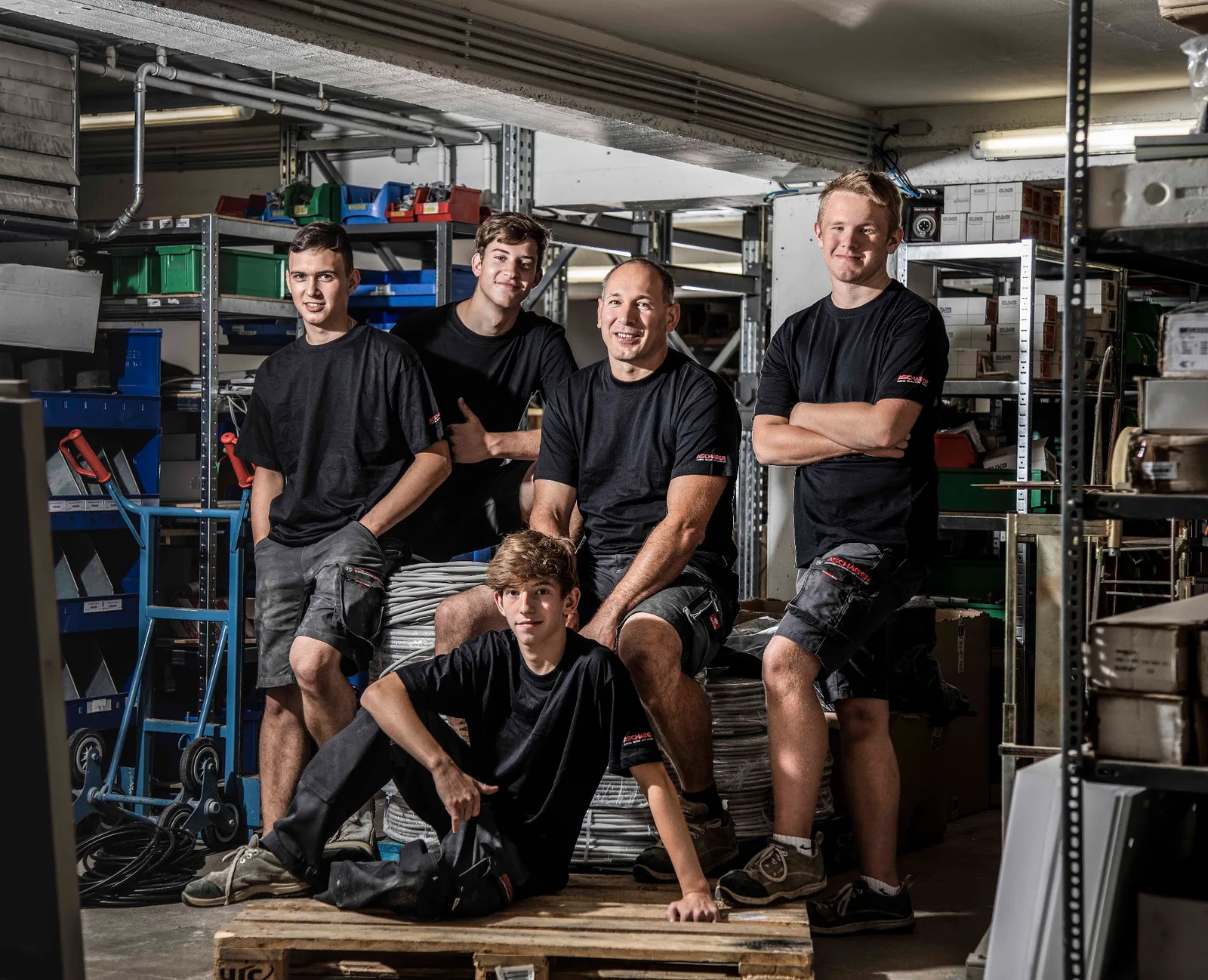 Fünf Mitarbeiter in schwarzen Firmen-T-Shirts sitzen im Lager auf Kabeltrommeln und einer Palette